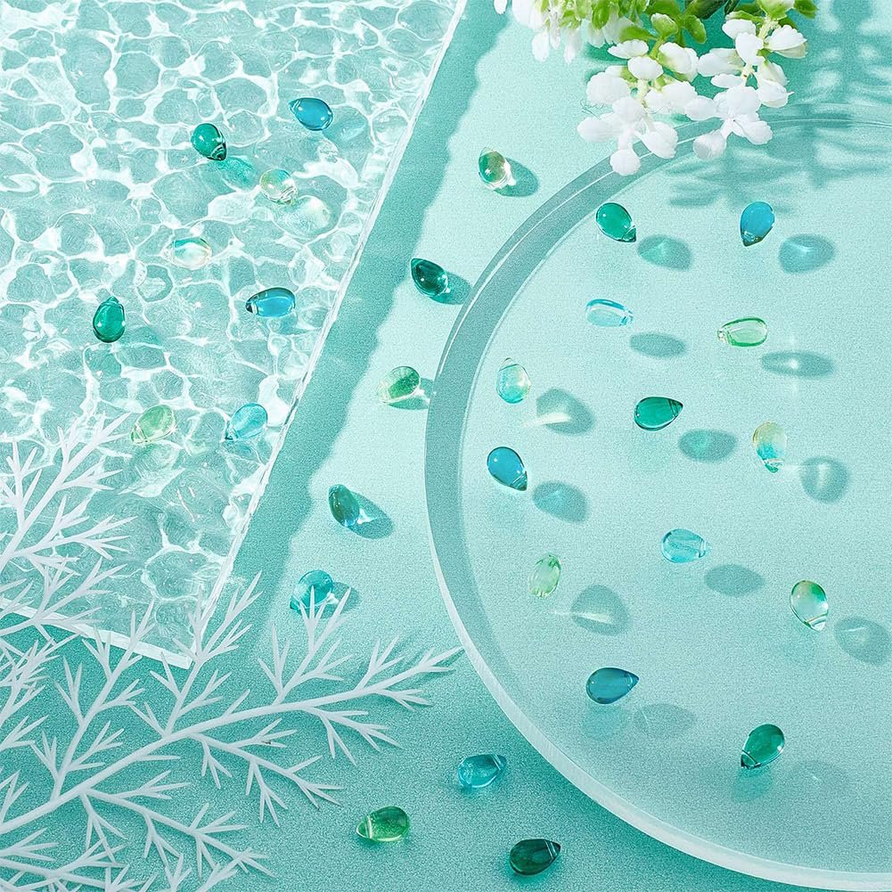 Teardrop glass beads in blue and green scattered on a teal surface with decorative white branches and flowers.