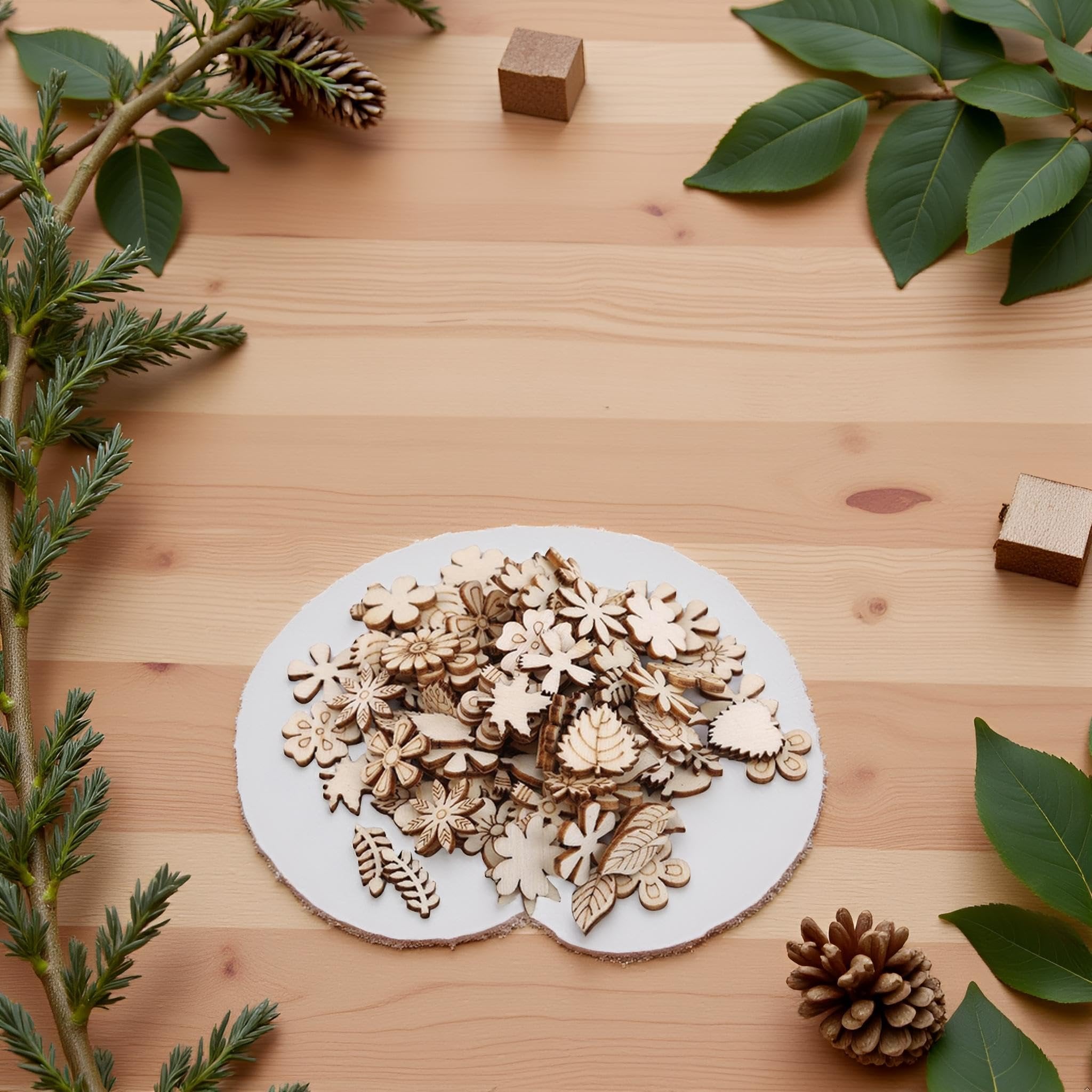 Pile of unfinished wooden flower and leaf craft cutouts on a white circular mat, surrounded by greenery and pinecones.