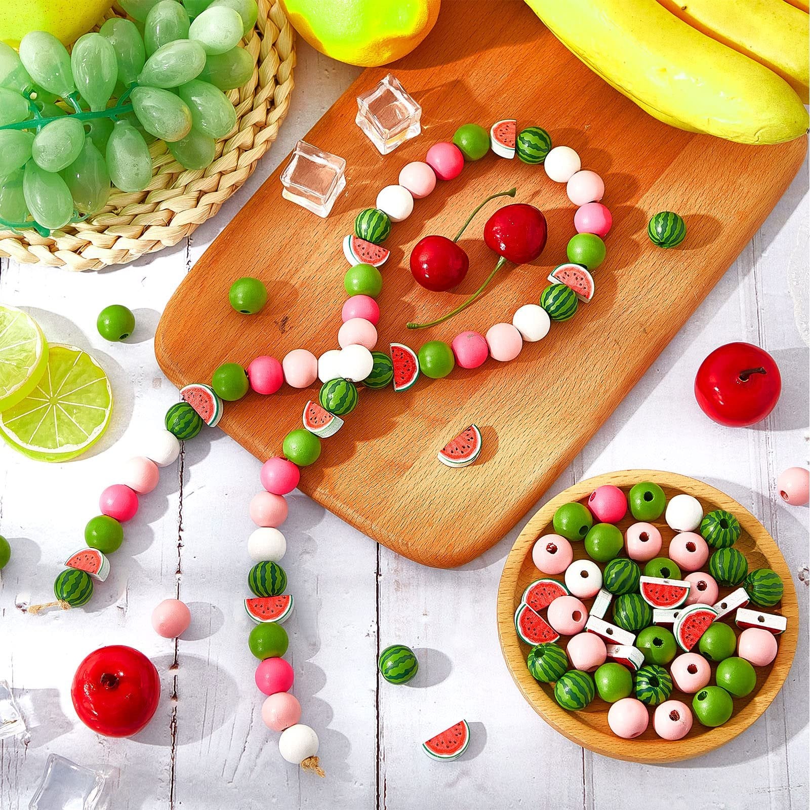 Assortment of watermelon and solid colored wooden beads arranged on a wooden cutting board and in a wooden bowl, with decorative fruit and ice cubes.