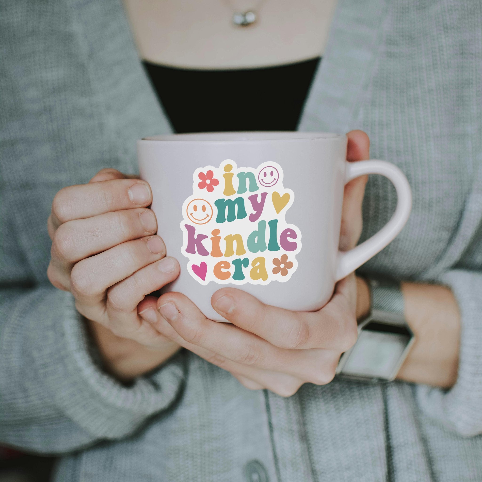 Person holding a mug with a colorful sticker that says 'in my kindle era' with smiley faces and hearts.