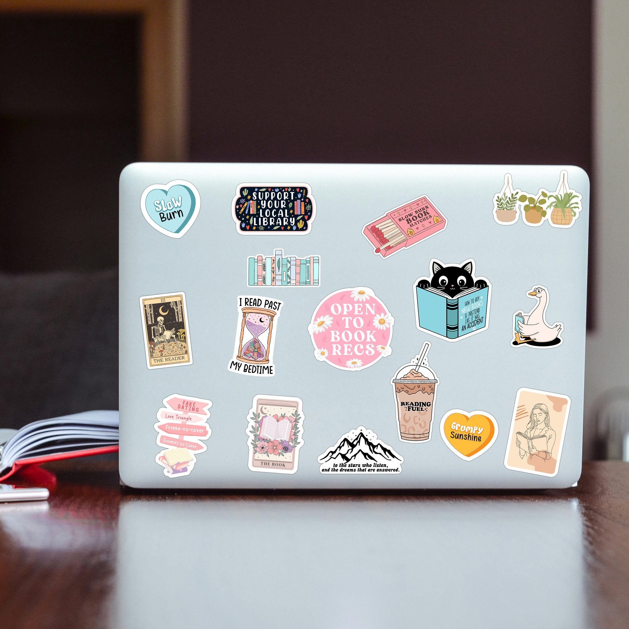Assortment of book-themed vinyl stickers applied to a laptop lid, featuring various designs like books, a cat, a duck, and quotes.