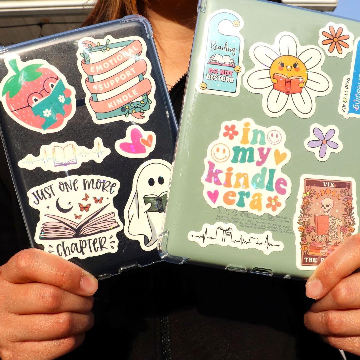 Two e-readers decorated with book-themed vinyl stickers, held by a person. One e-reader has stickers like a strawberry reading and 'Emotional Support Kindle'.