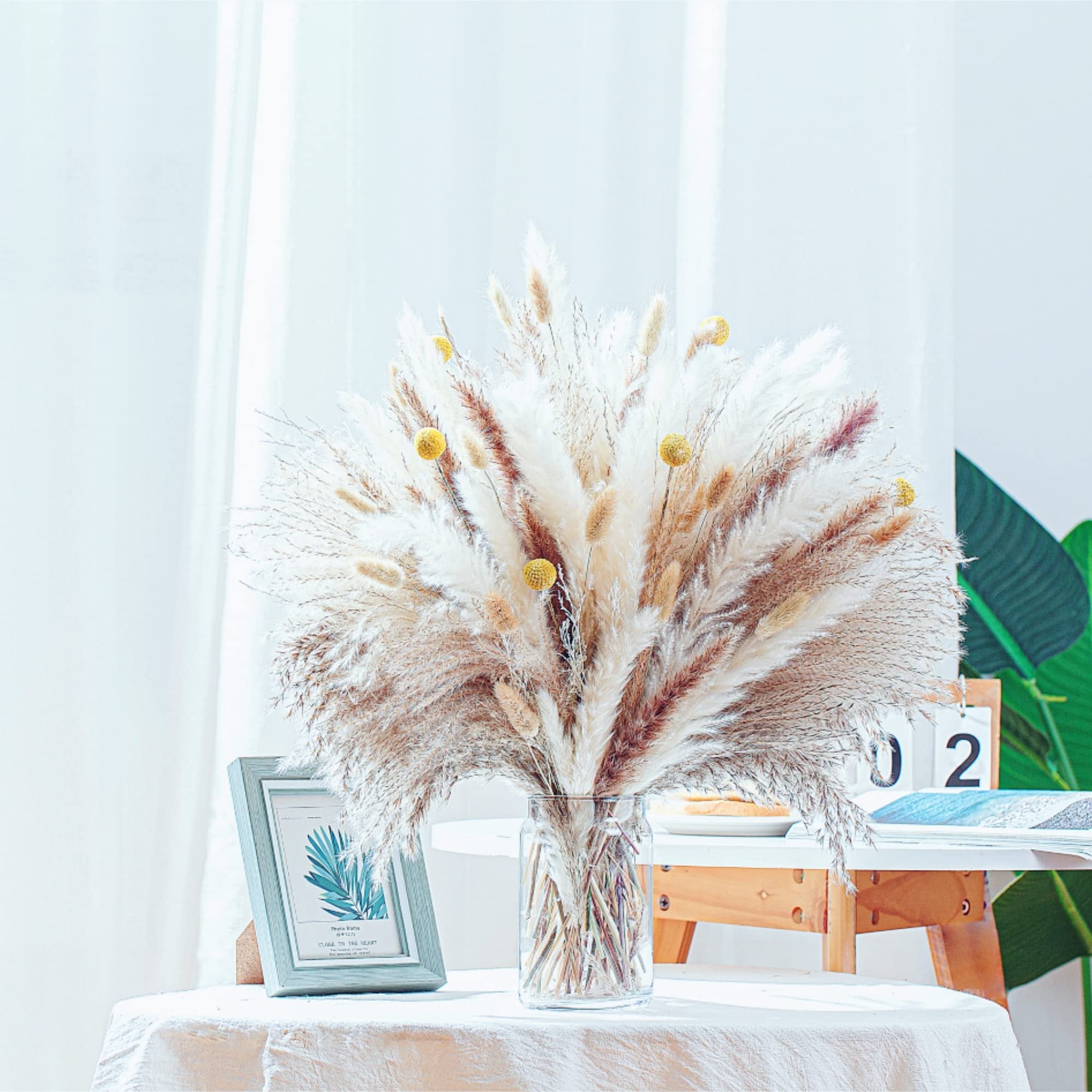 Bouquet of dried pampas grass and small yellow flowers in a glass vase on a table, with a framed picture and calendar in the background.