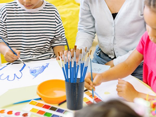 Children and adult painting with watercolor set and paint brushes in a classroom setting