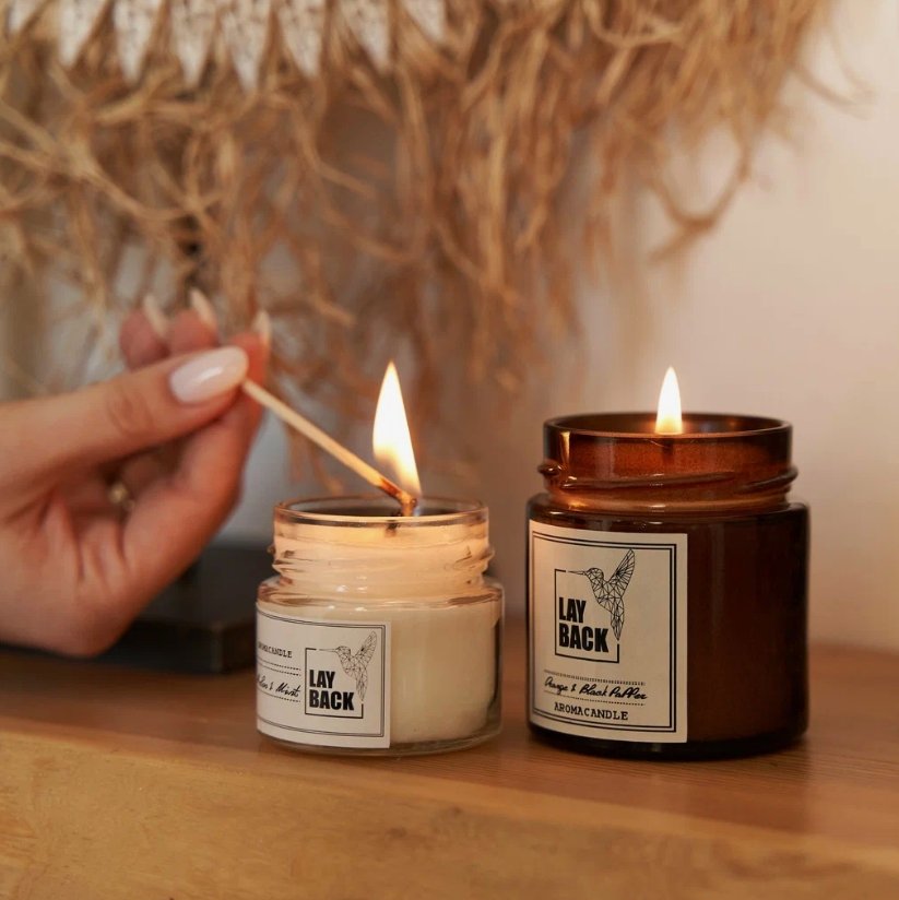 Two scented candles on a wooden surface, one being lit by a match. The candle on the right is in a brown glass jar.