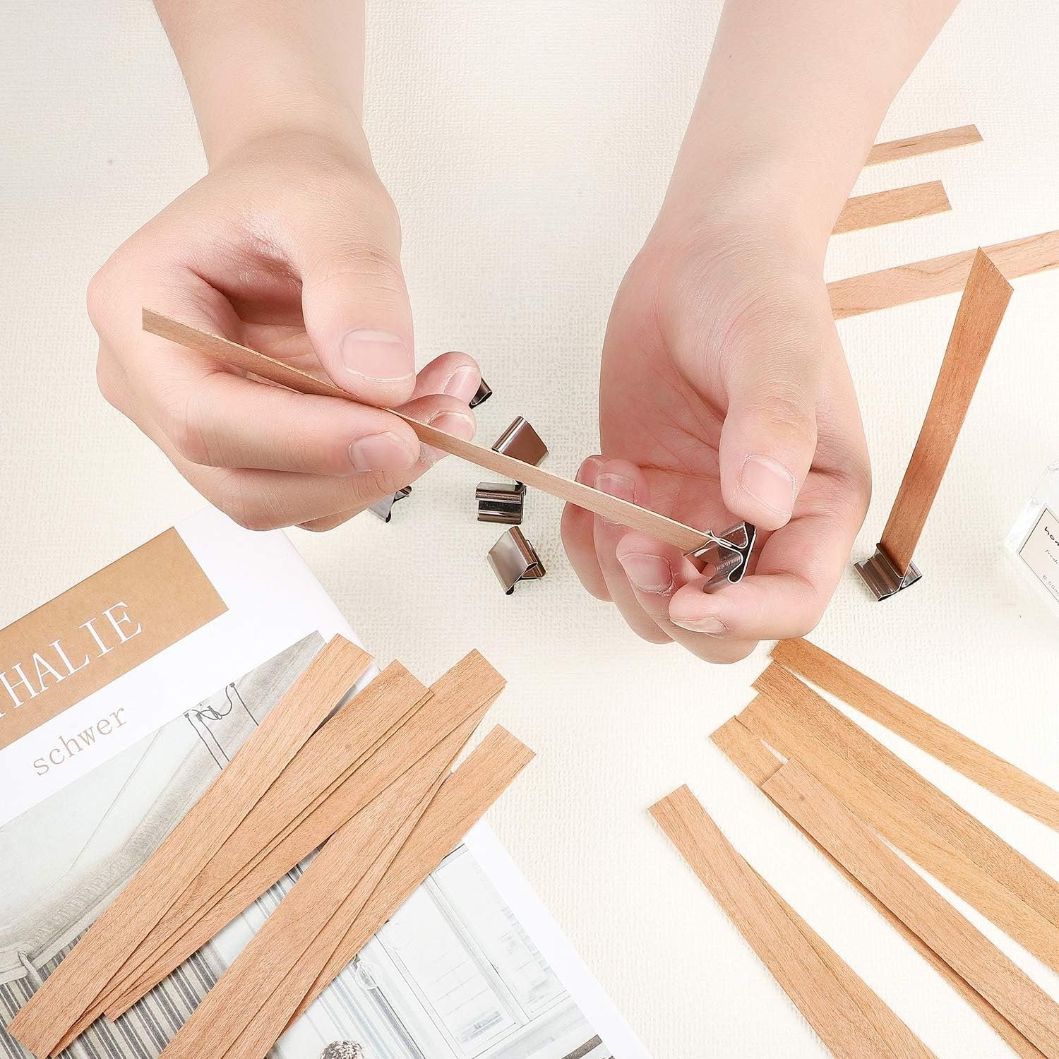 Hands assembling wooden candle wicks with metal clips and stickers, surrounded by wicks and a book.