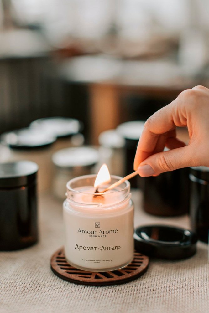 Hand lighting a candle with a match, showing the glass jar with label and wooden wick on a coaster.