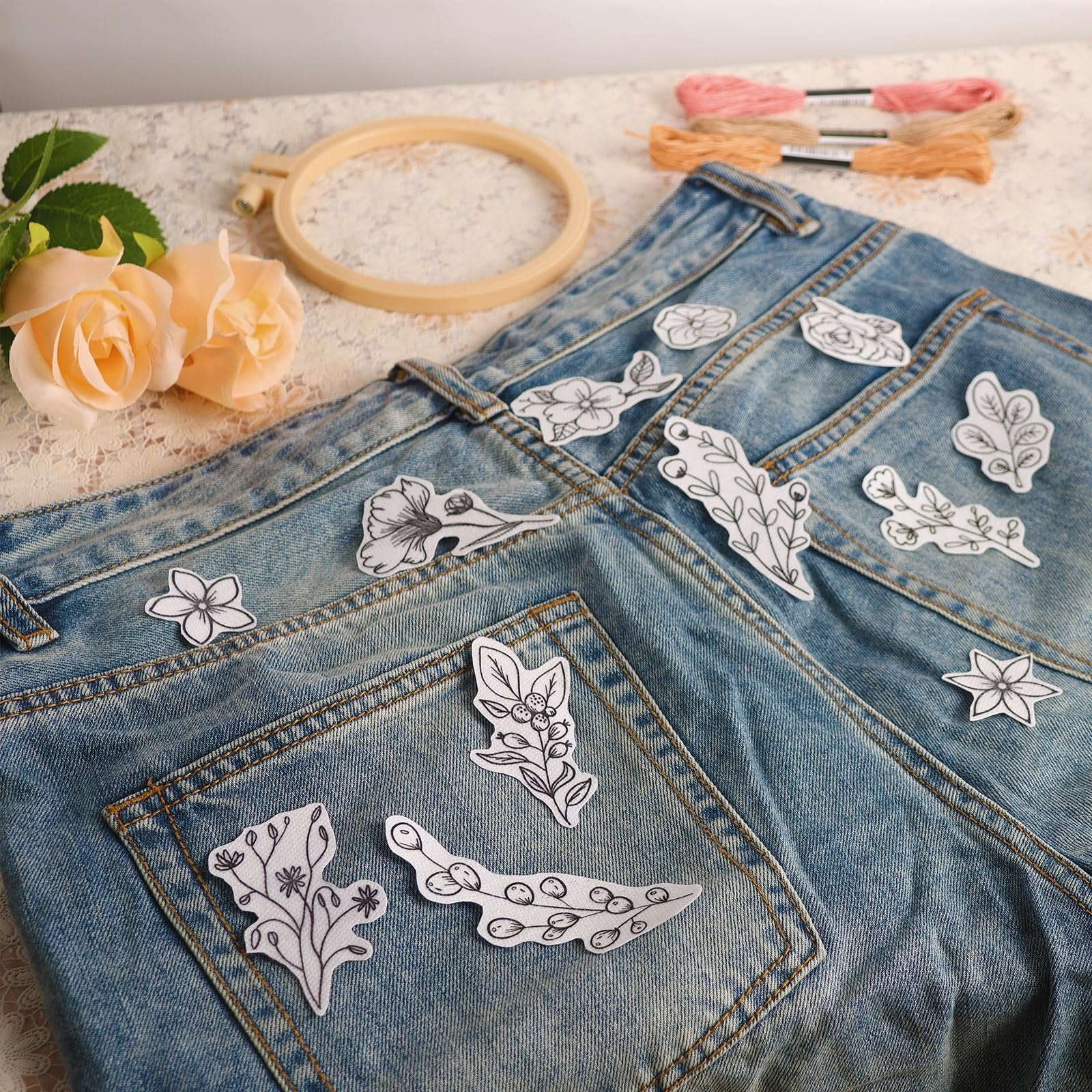 Water soluble embroidery stabilizer patterns with floral and leaf designs adhered to denim jeans, with embroidery hoop and floss in background.