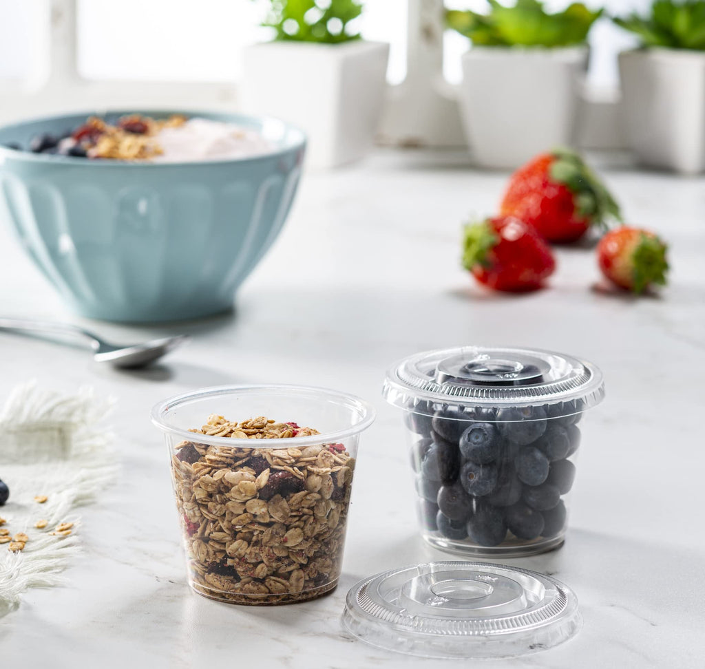 Two clear plastic portion cups with lids, one filled with granola and berries, the other with blueberries, next to a bowl of yogurt and fruit.