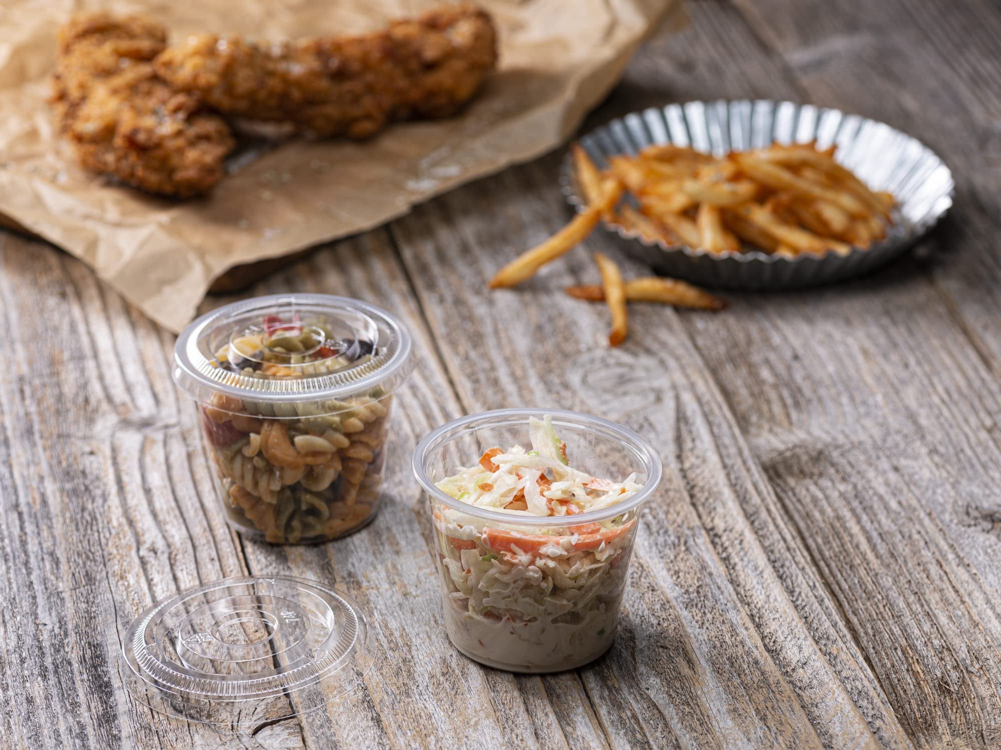 Two clear plastic portion cups filled with pasta salad and coleslaw, with fried chicken and french fries in the background on a wooden table.