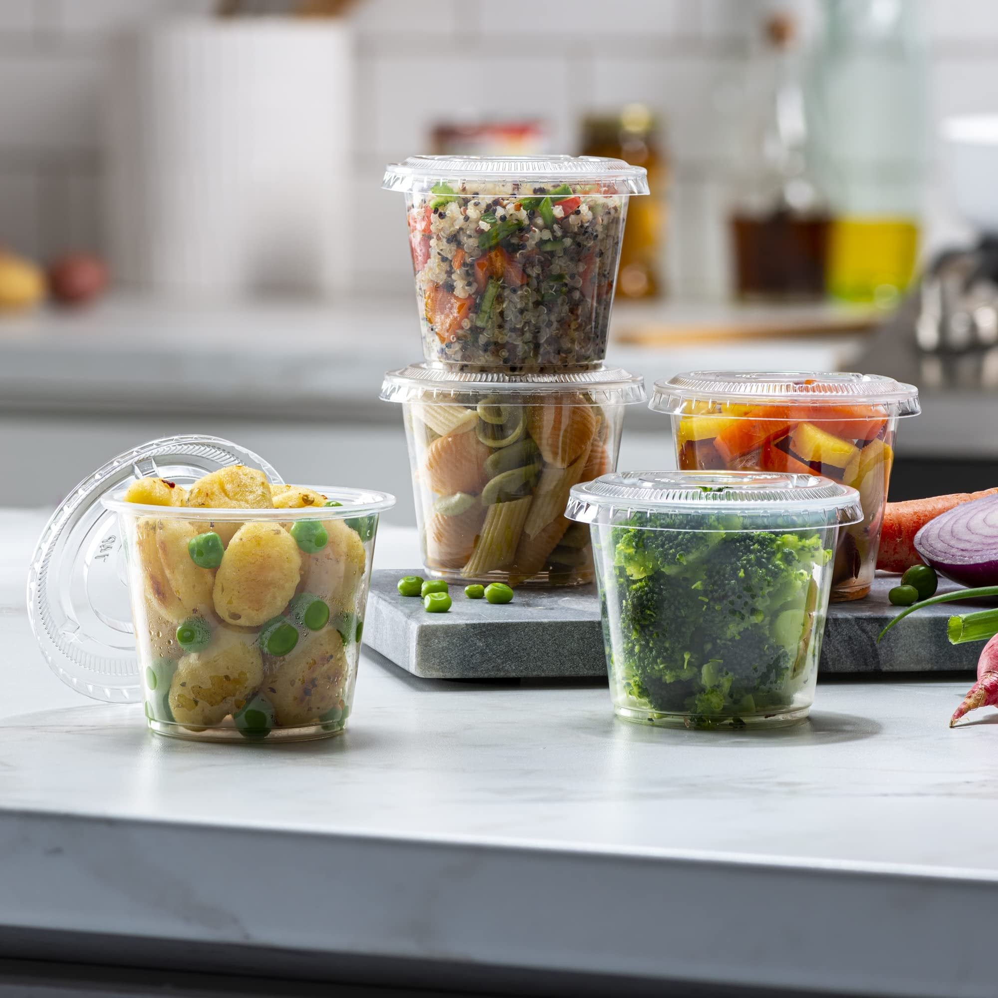 Four clear plastic portion cups filled with food, stacked and arranged on a countertop with vegetables.