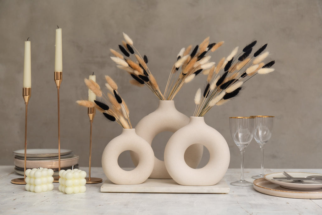 Dried bunny tail grass in black, white, and natural brown arranged in modern beige vases on a table with candles and plates.