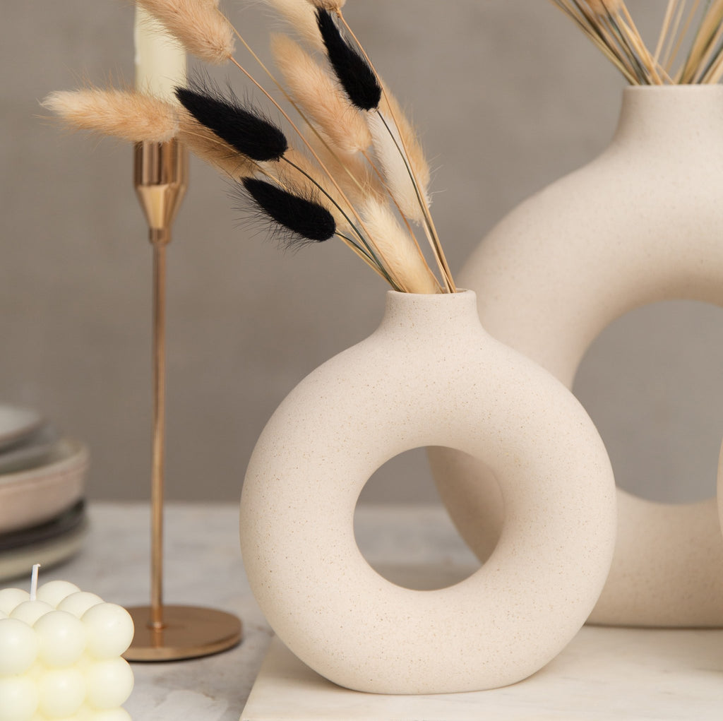 Donut-shaped ceramic vase with dried bunny tail grass and black accents, next to a gold candlestick and bubble candle.