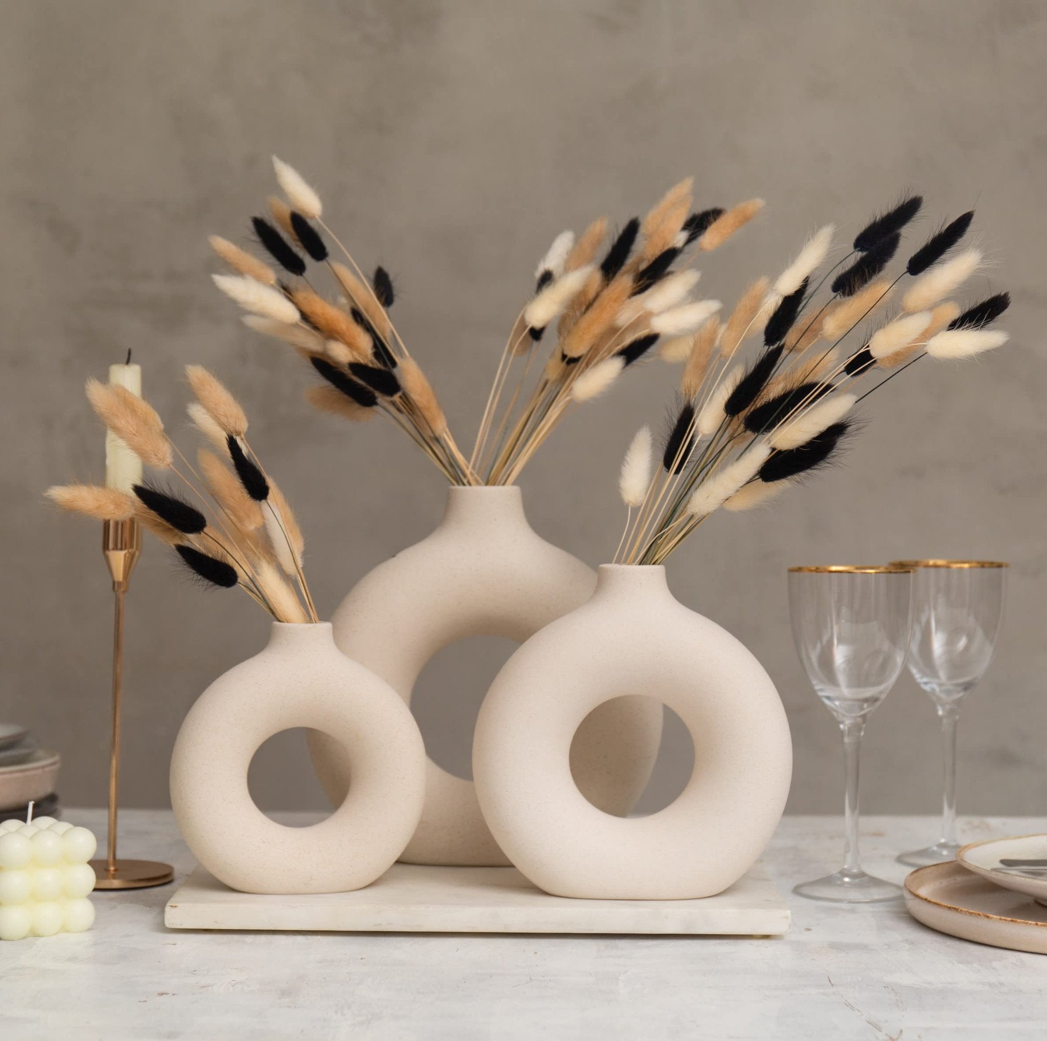 Three modern beige vases with dried bunny tail grass in black, white, and natural brown, arranged on a table with a candle and wine glasses.