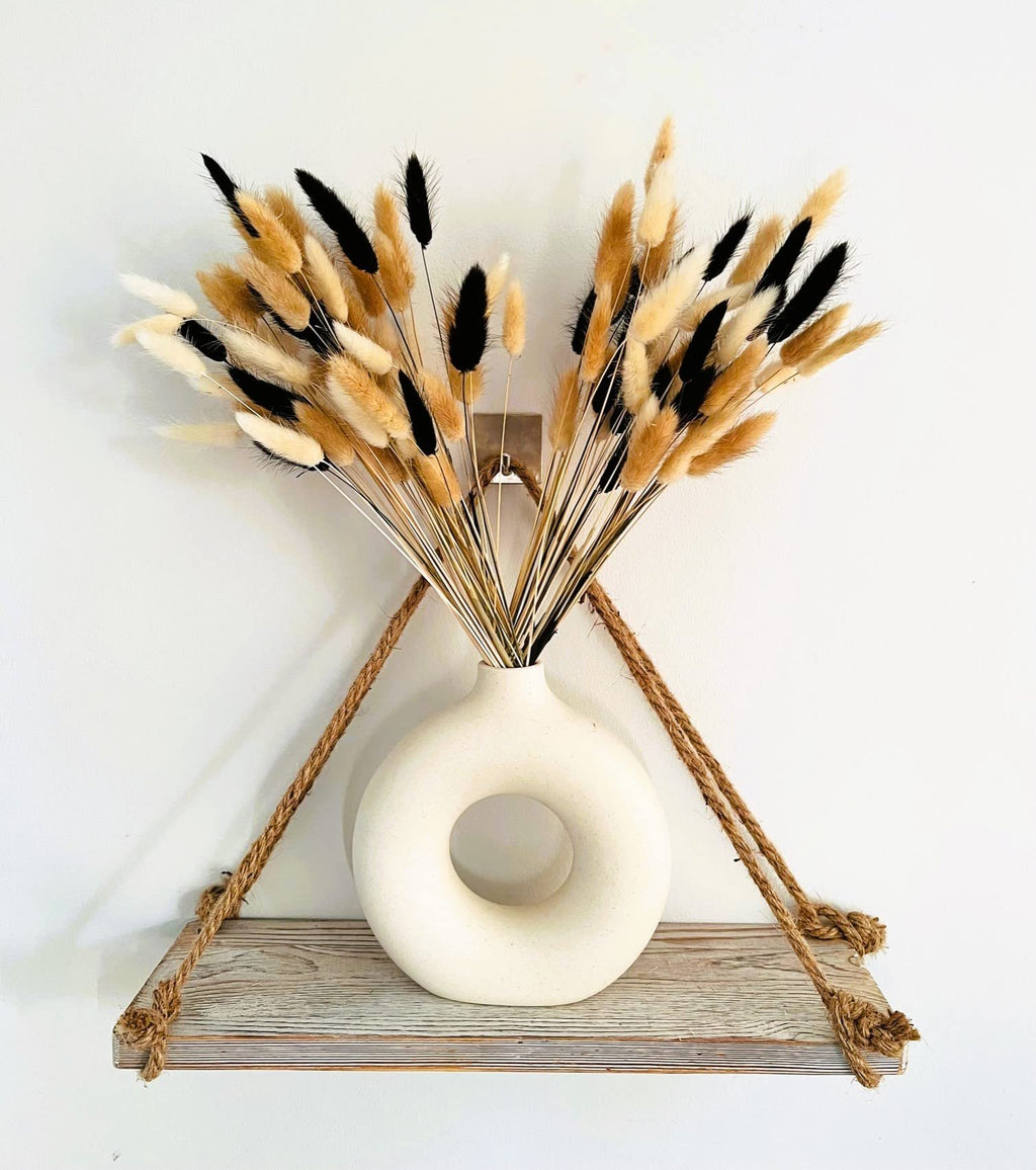 Bunny tails pampas grass dried flowers in a white donut vase on a wooden shelf with rope accents