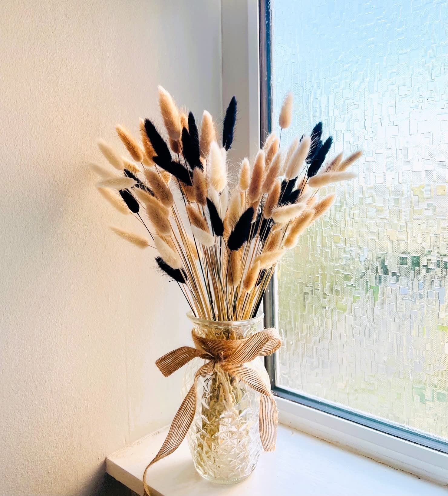 Bunny tail dried flowers in a glass vase with a burlap ribbon, placed on a windowsill.