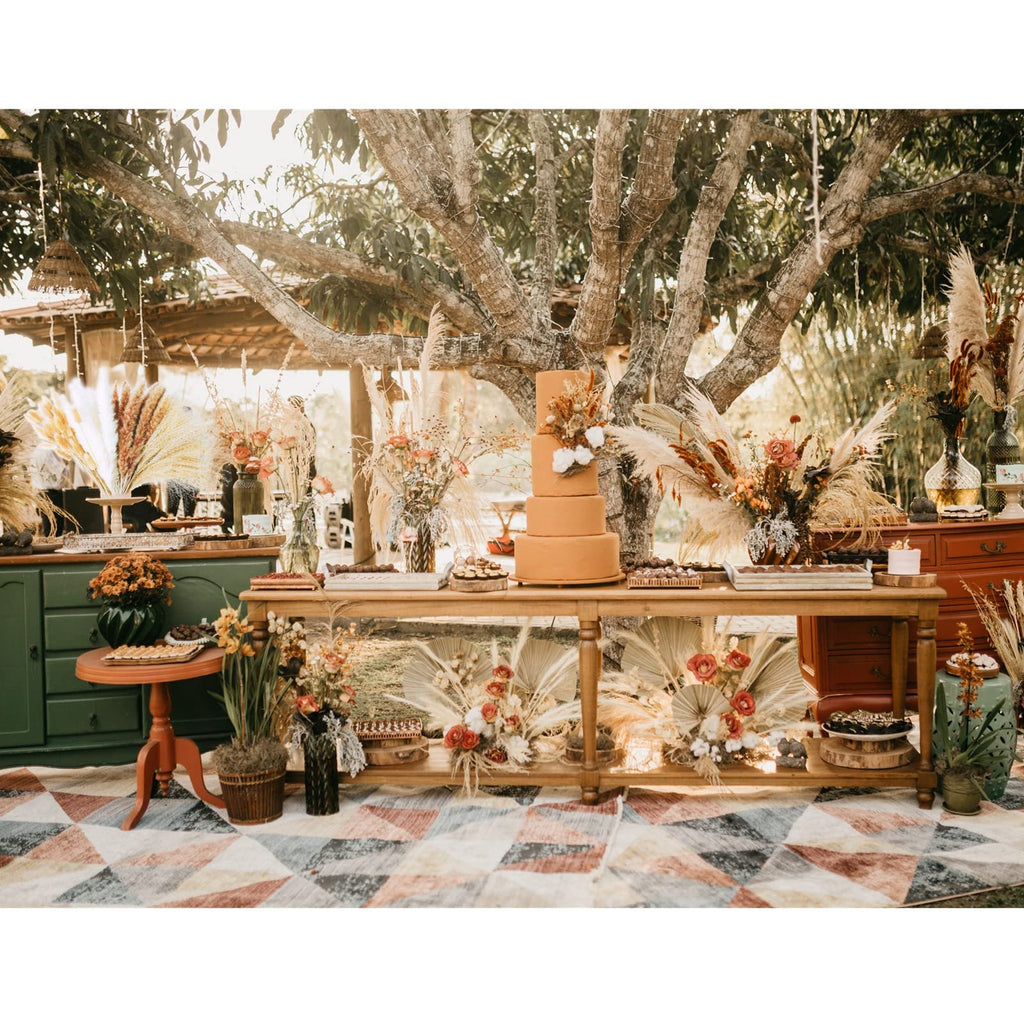 Outdoor dessert table with a tiered cake, floral arrangements, and dried pampas grass under a tree.