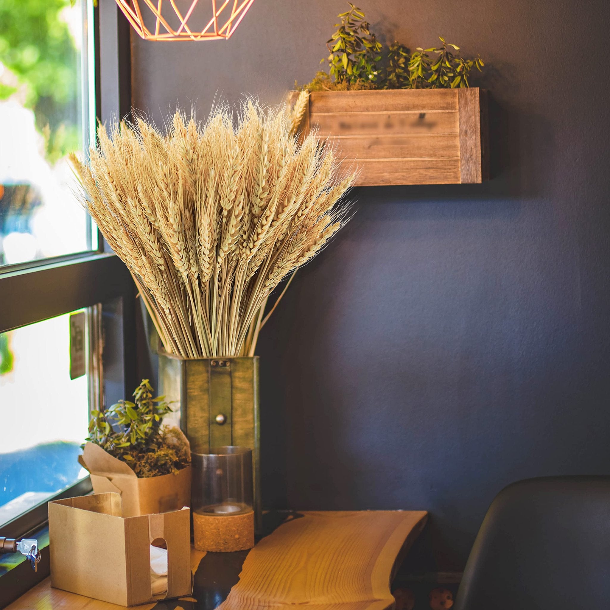 Bundle of dried wheat stalks in a rustic green vase next to a wooden planter with greenery.