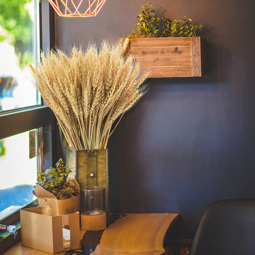 Bundle of dried wheat stalks in a rustic green vase next to a wooden planter with greenery.