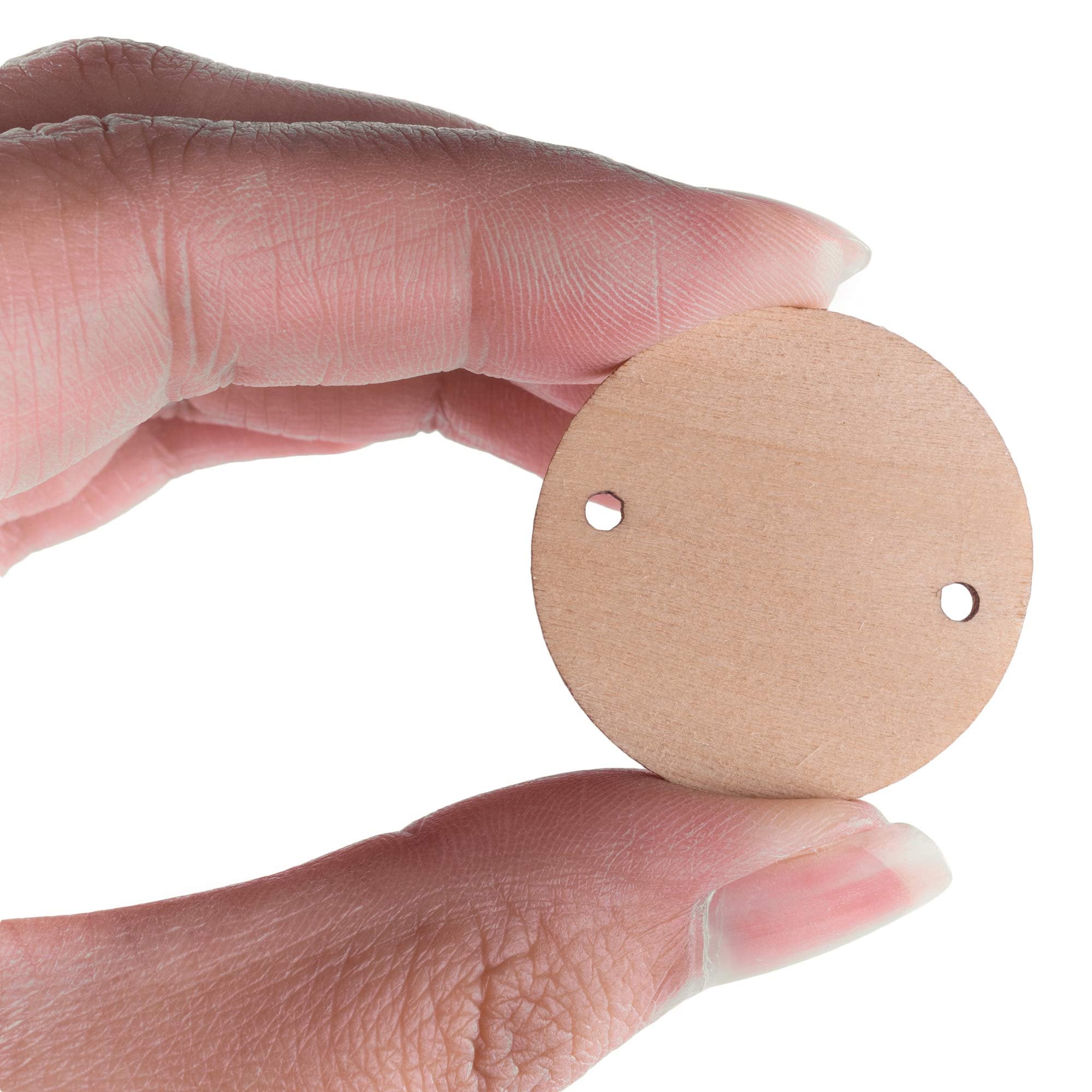Fingers holding a 1.2-inch unfinished wooden round disc with two pre-drilled holes for crafts.
