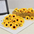 Pile of 100 yellow artificial sunflower heads scattered on white paper with a wooden crate in the background.