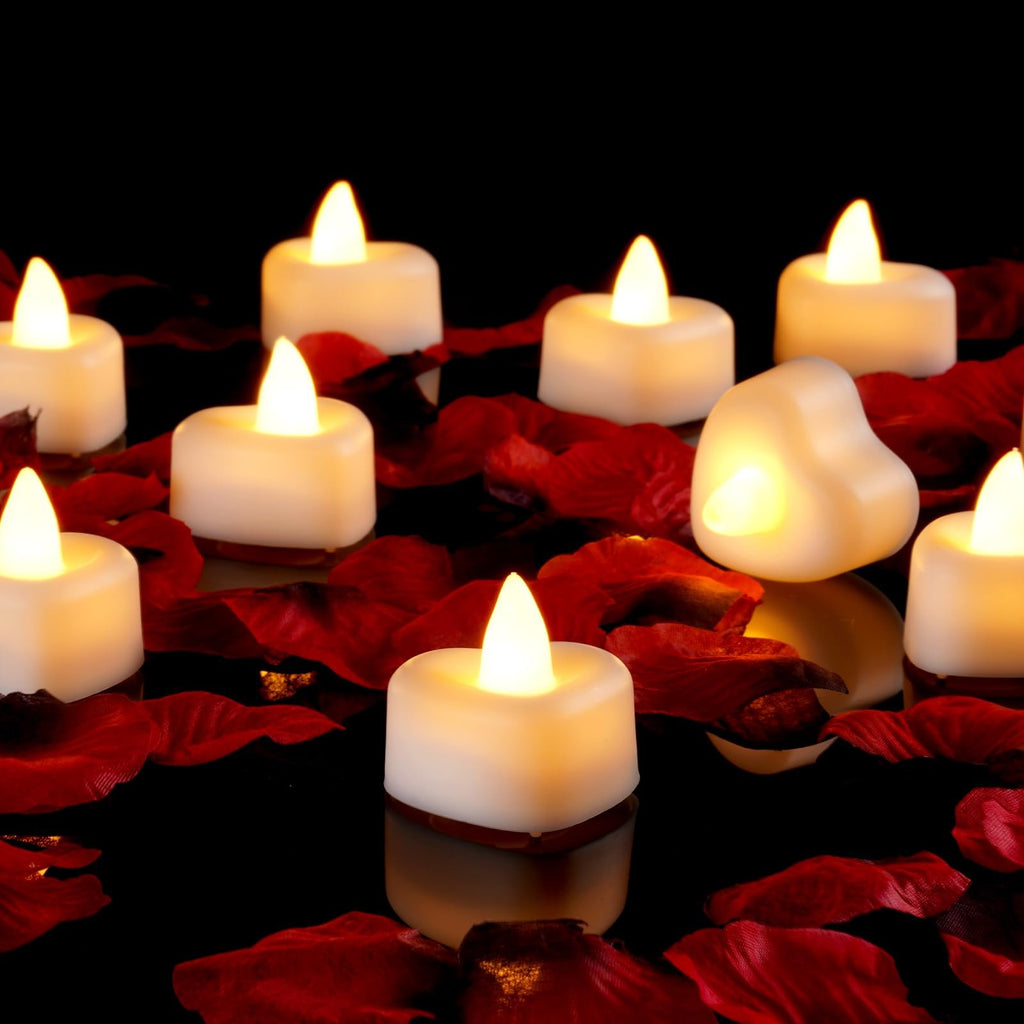 Heart-shaped LED tea lights scattered amongst red rose petals on a reflective black surface.