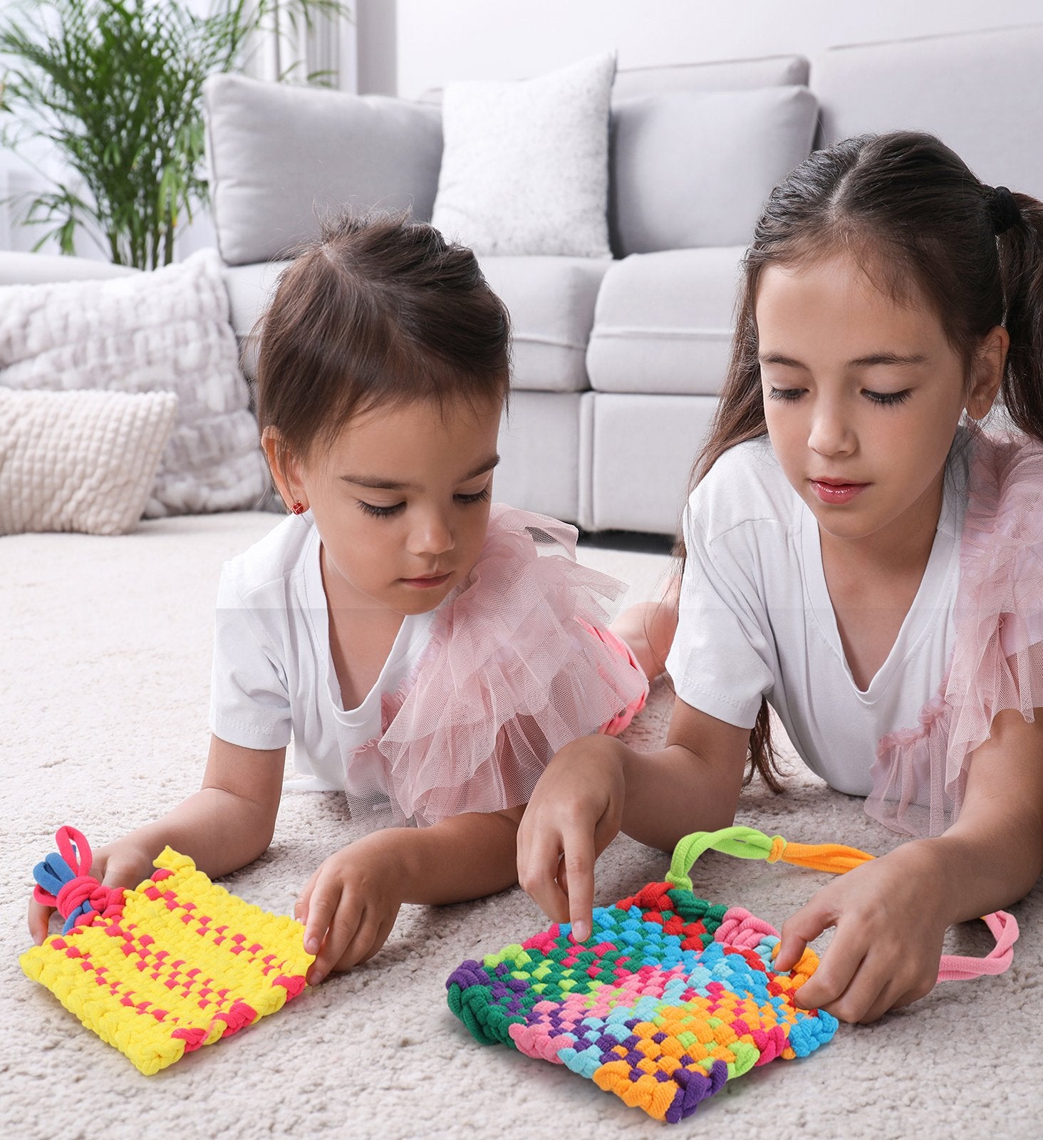 Two young girls lying on a carpet, weaving colorful fabric squares with a loom kit.