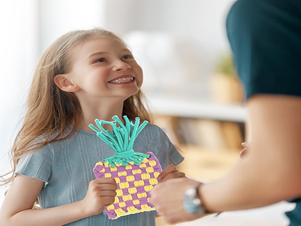 Smiling girl holding a woven craft project with teal yarn strands sticking out, receiving it from an adult.