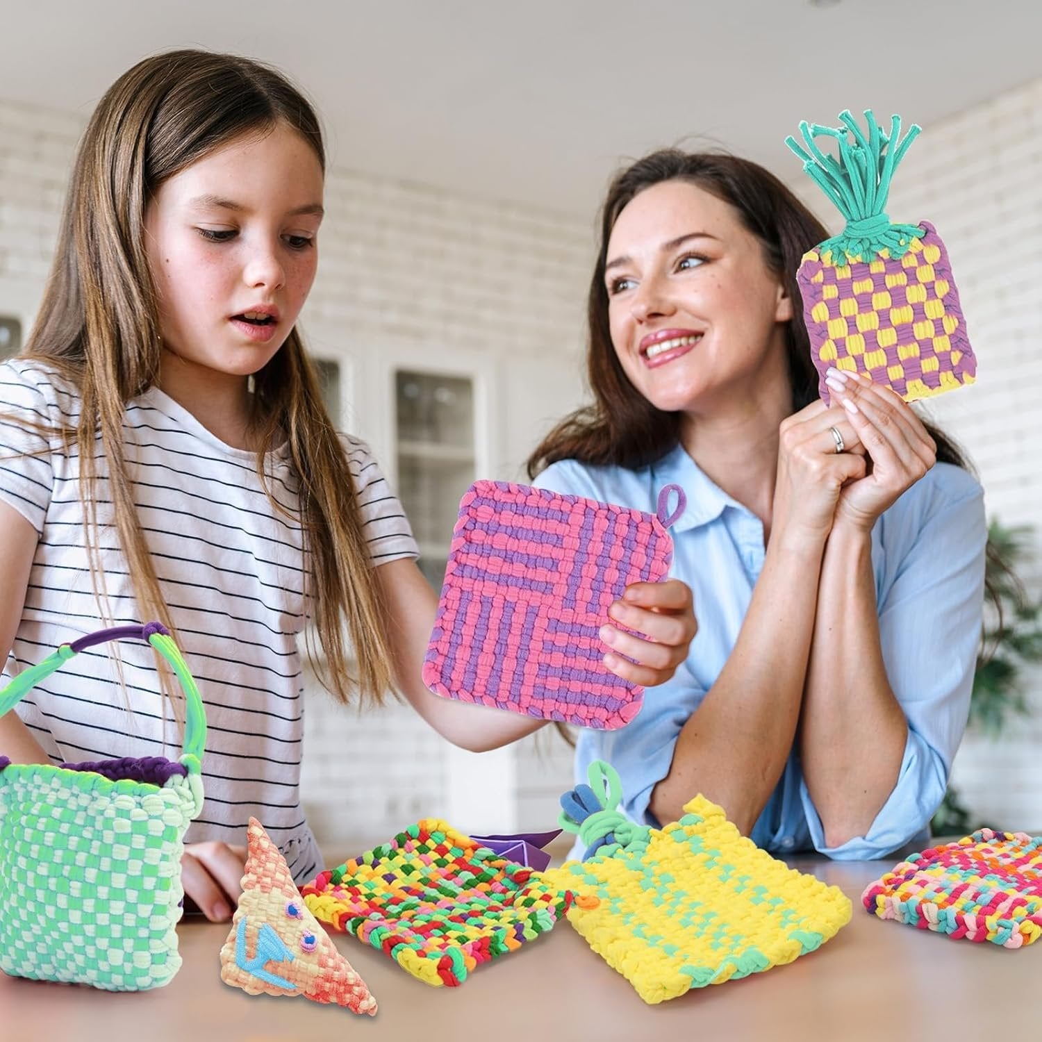 Woman and girl holding colorful woven crafts made with loom loops, with more crafts on table.