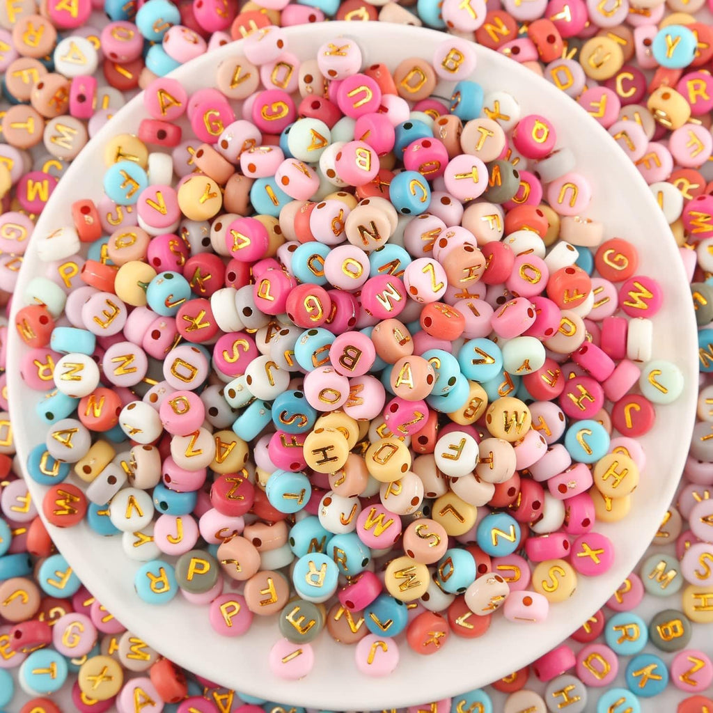Top-down view of colorful round acrylic letter beads with gold letters, spilling out of a white bowl.