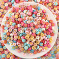 Top-down view of colorful round acrylic letter beads with gold letters, spilling out of a white bowl.