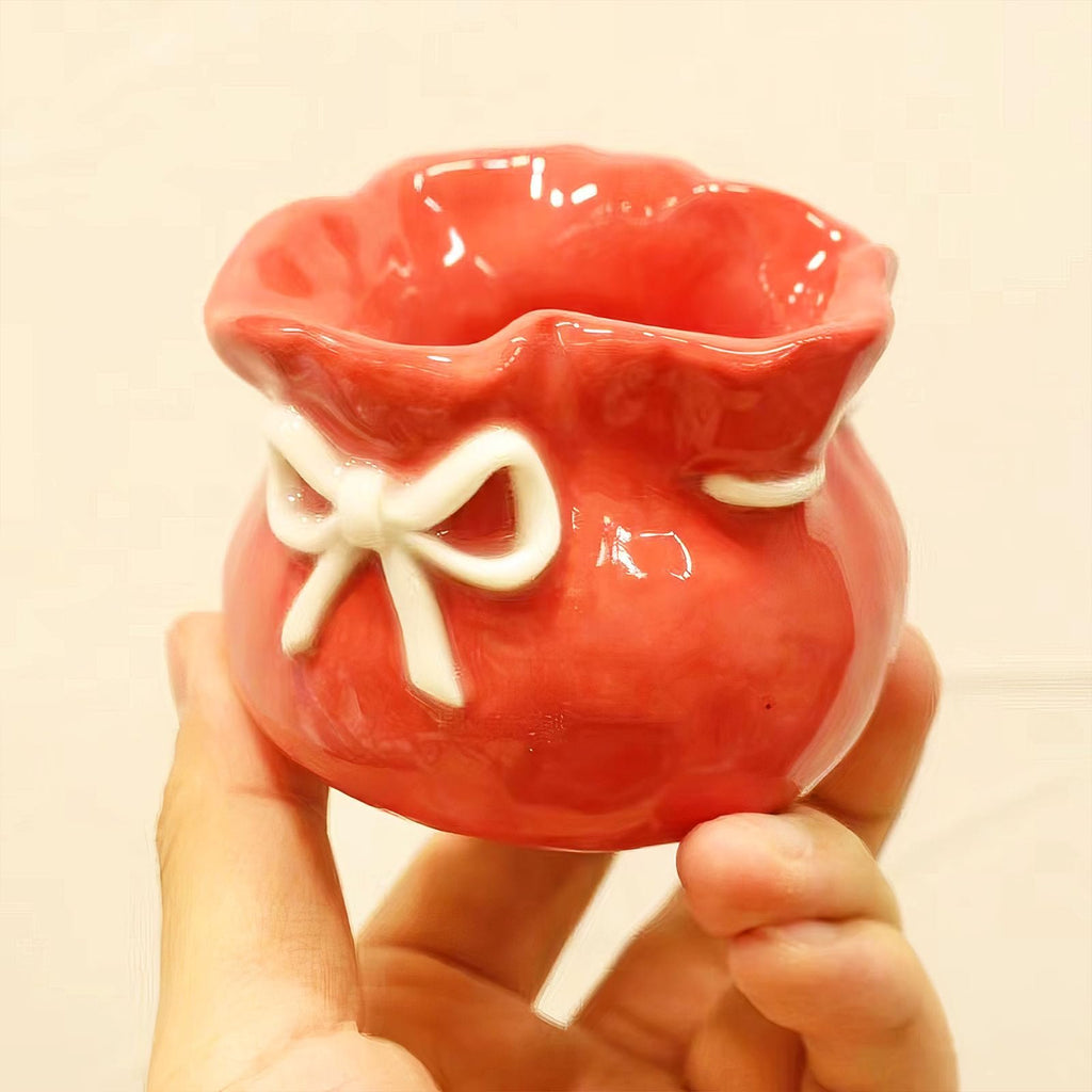 Hand holding a red ceramic pot shaped like a gift bag with a white bow detail.
