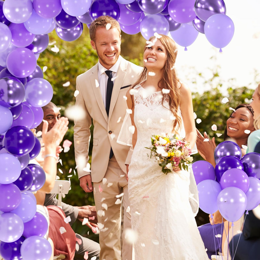 Bride and groom smiling at outdoor wedding ceremony with purple balloons and falling petals