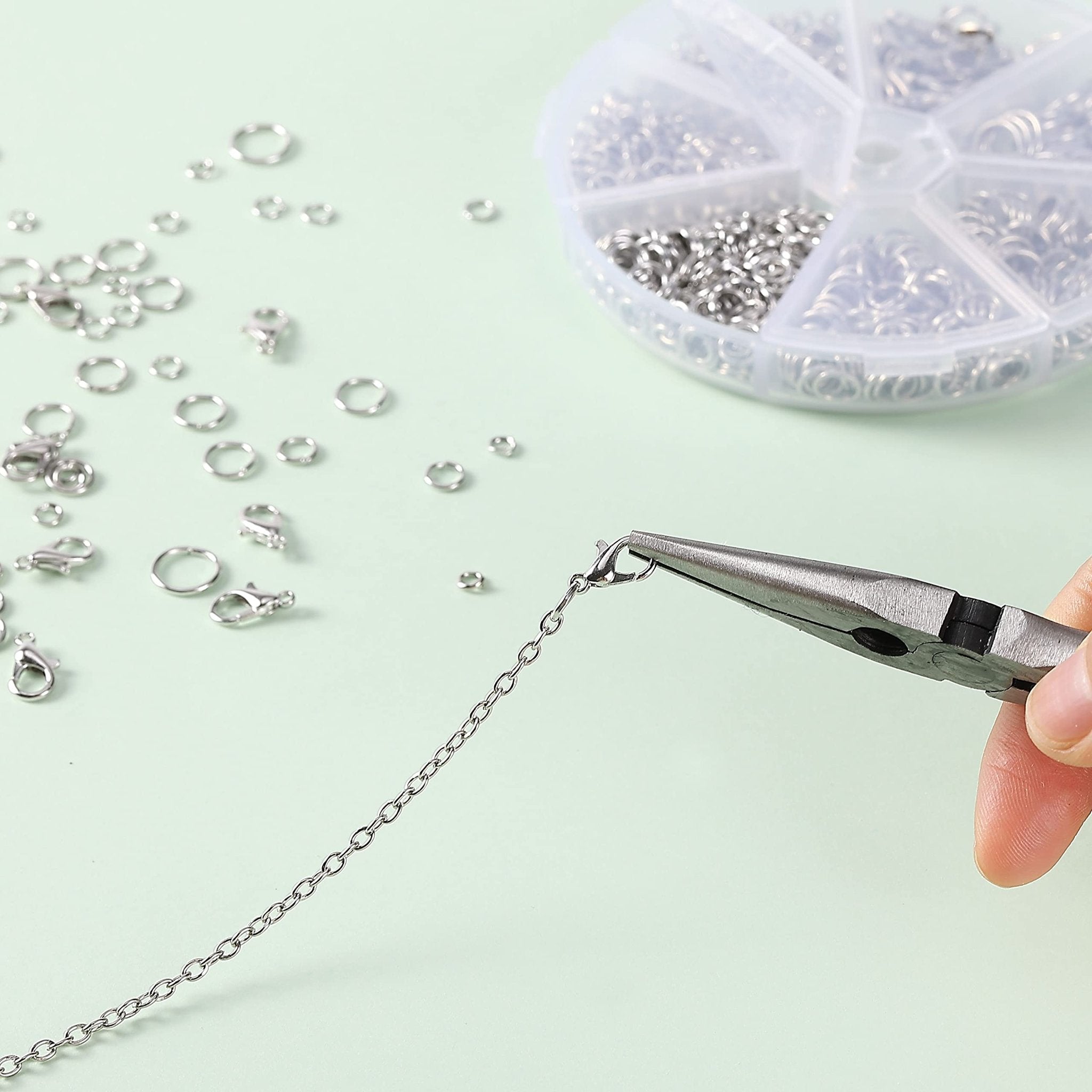 Close-up of a hand using pliers to attach a lobster clasp to a silver chain, with jump rings and a storage case scattered on a light green background.