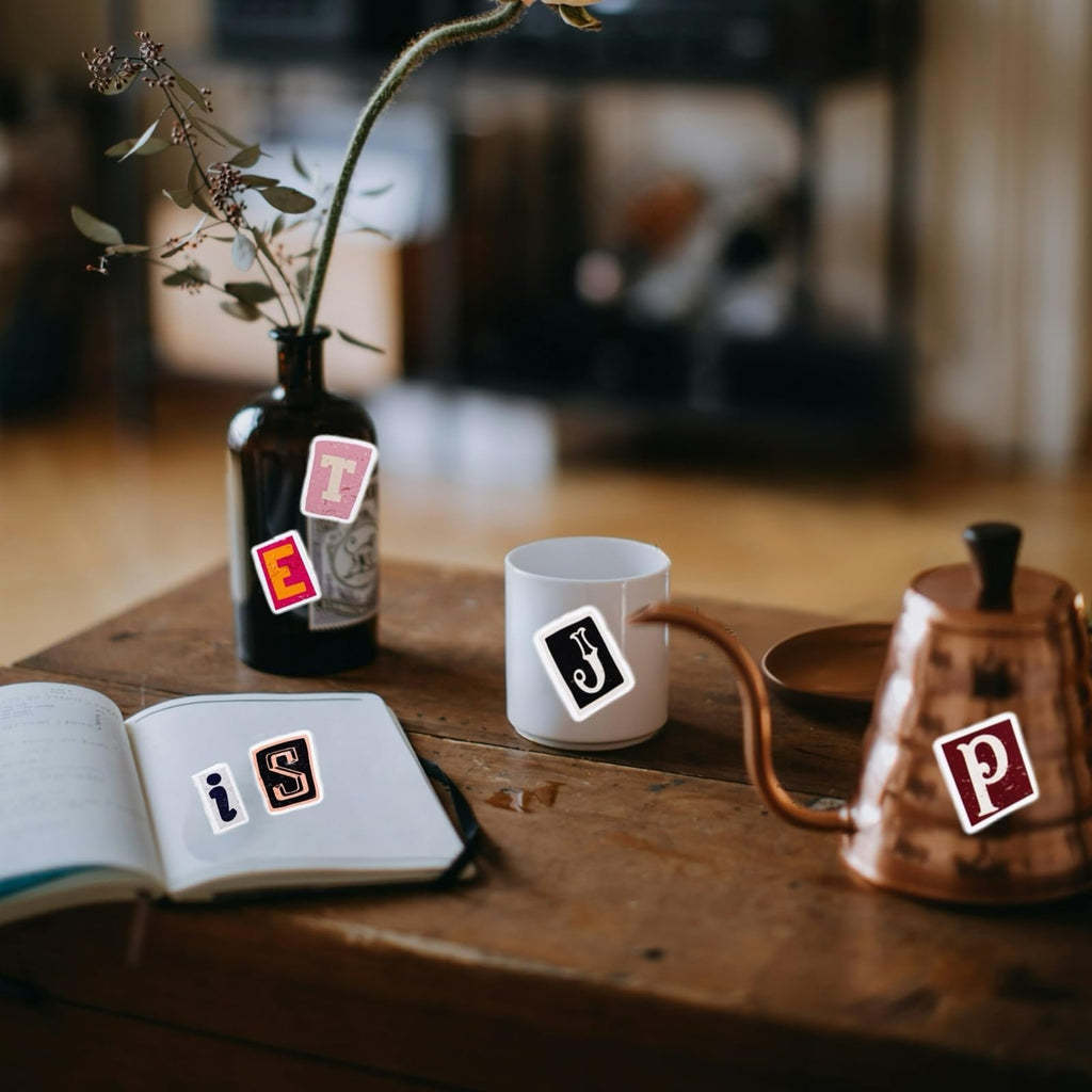 Vintage alphabet stickers on a notebook, vase, and mug on a wooden table.