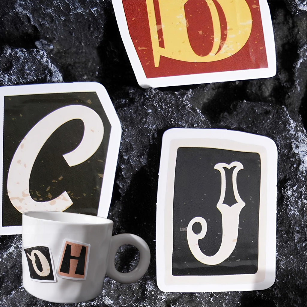 Vintage alphabet stickers C, D, J, O, H on a white mug and dark textured surface