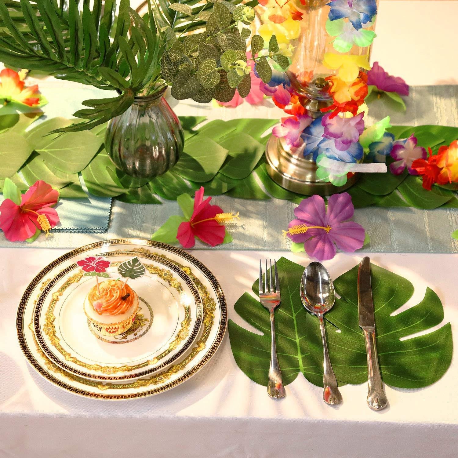 Tropical table setting with a cupcake on stacked plates, silverware on large leaves, and floral decorations.