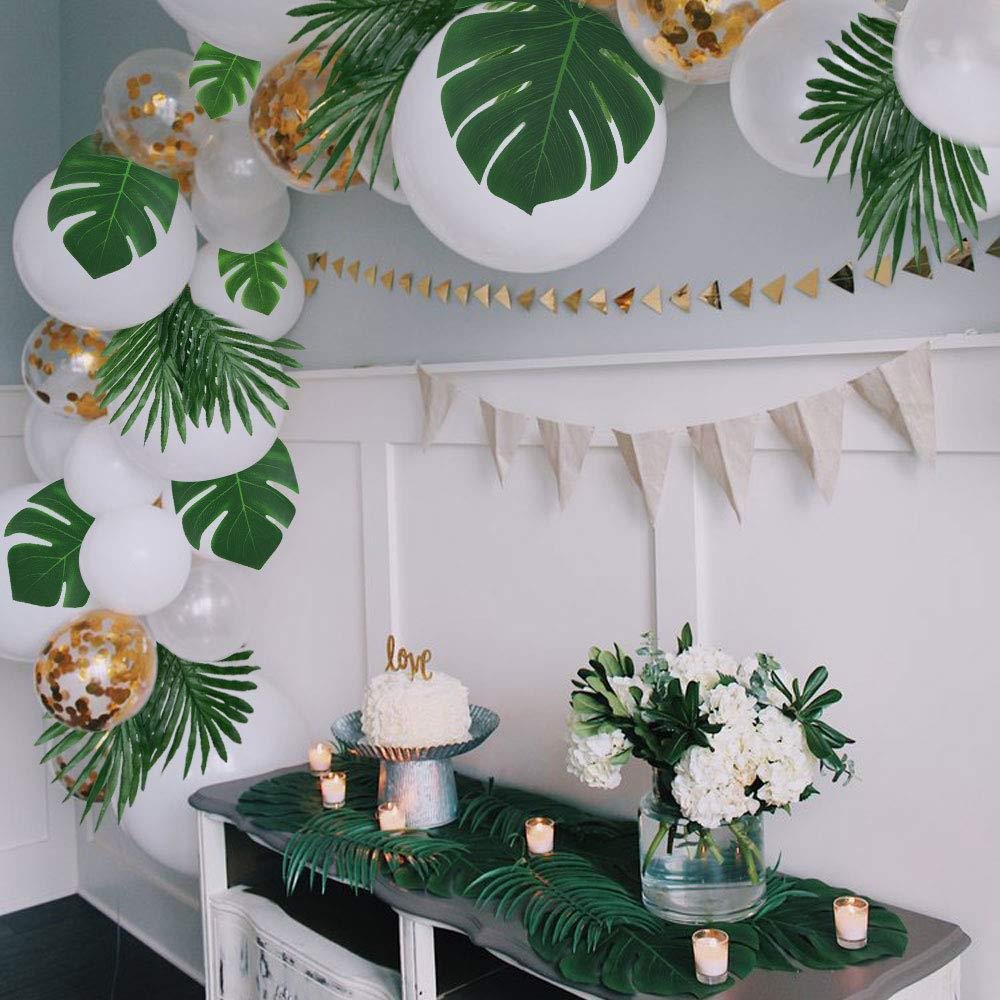 Tropical party decor with artificial palm leaves and white balloons, cake, and flowers on a table.