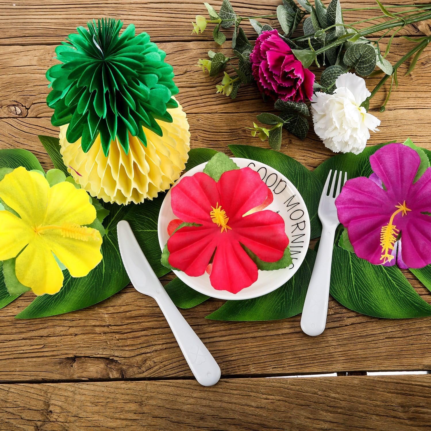 Tropical party decor set with pineapple honeycomb decorations, hibiscus flowers, and leaves arranged on a wooden table with a plate and cutlery.