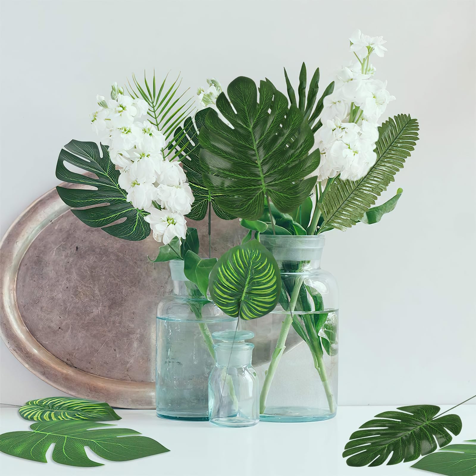 Artificial tropical leaves and white flowers arranged in glass vases on a shelf with a decorative tray
