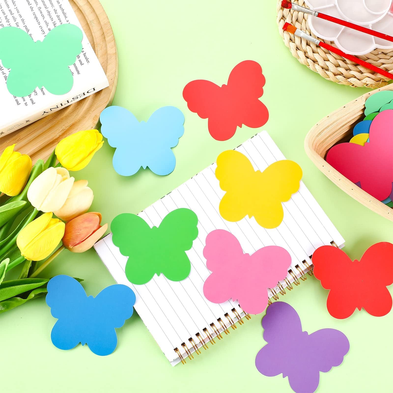 Colorful butterfly paper cutouts scattered on a notebook with tulips and art supplies in the background.