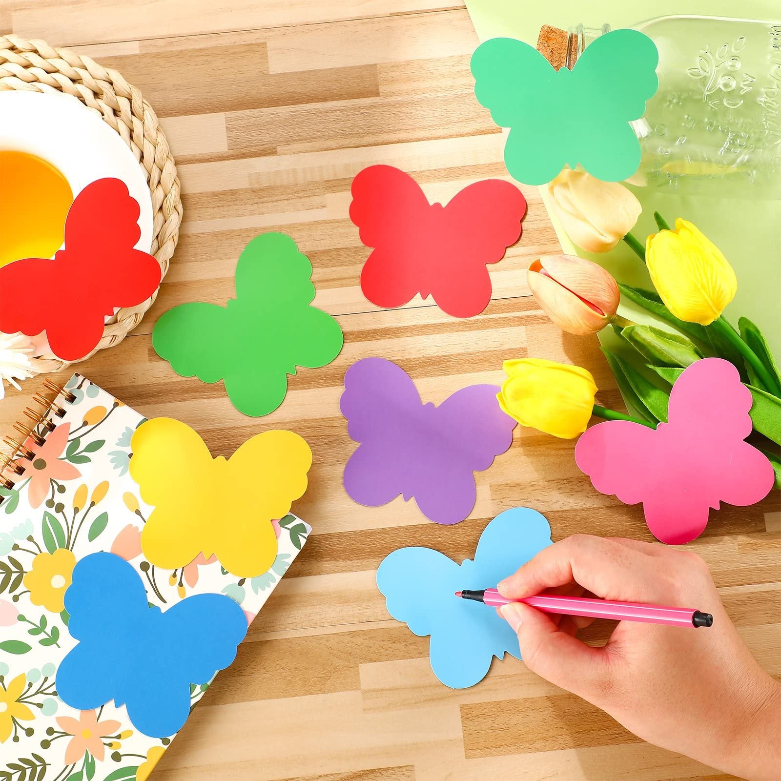Colorful butterfly paper cutouts scattered on a wooden surface with a notebook and tulips, a hand holding a pink marker over a blue cutout.