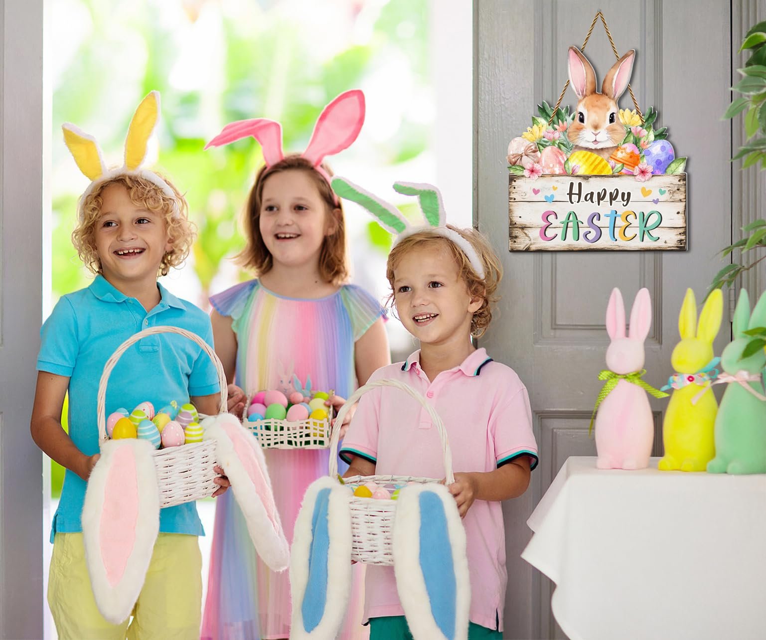 Three children wearing bunny ears and holding Easter baskets with eggs, a Happy Easter door sign hangs on a gray door.