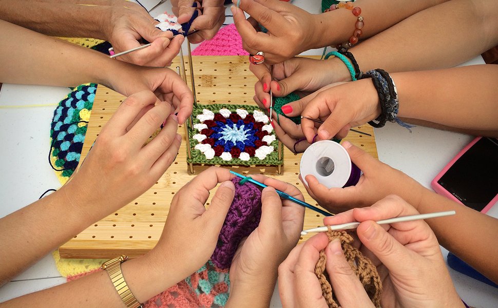 Multiple people crocheting granny squares on a bamboo blocking board with steel pins.