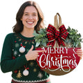 Woman in a festive sweater pointing to a round wooden Merry Christmas sign with a plaid bow and greenery