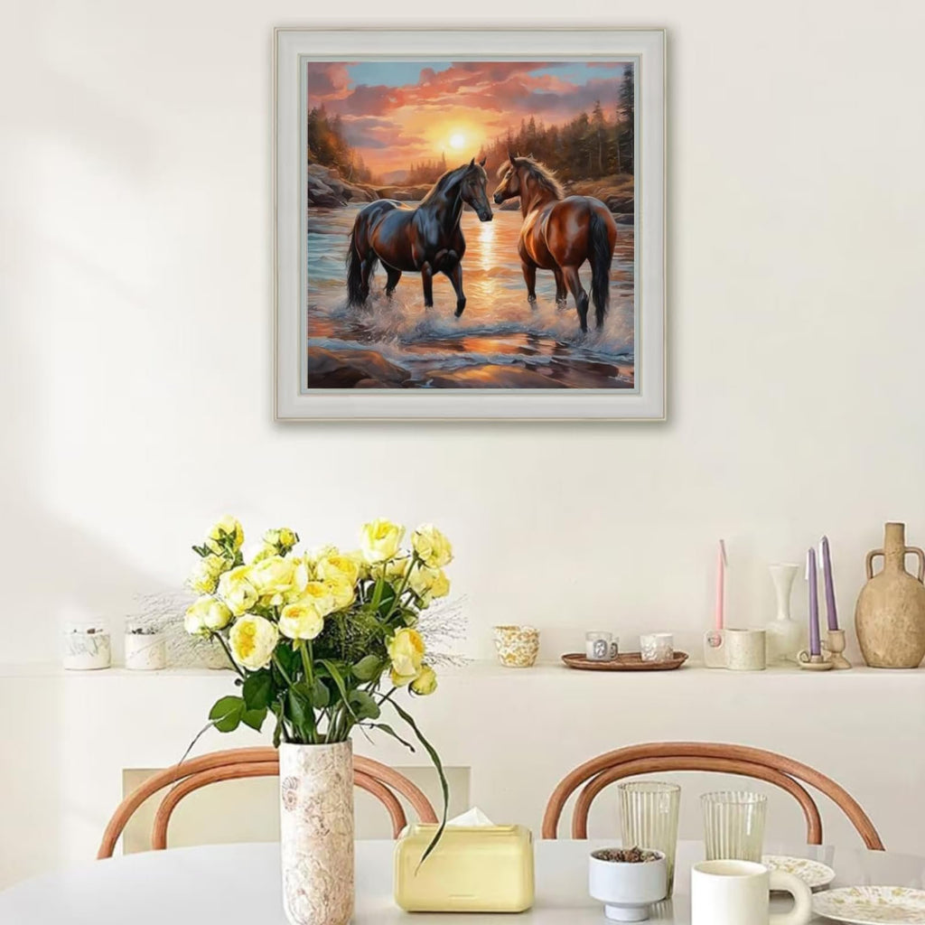 Diamond painting of two horses standing in a river at sunset, framed and hanging on a wall above a table with flowers.