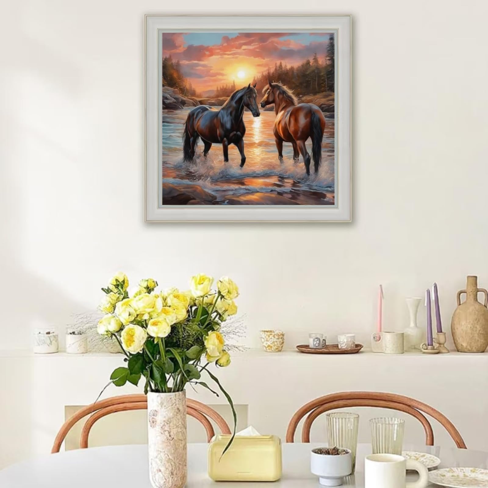 Diamond painting of two horses standing in a river at sunset, framed and hanging on a wall above a table with flowers.