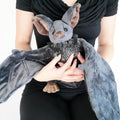 Person holding a gray plush bat with large wings and fluffy chest fur. The bat has brown inner ears and paws.