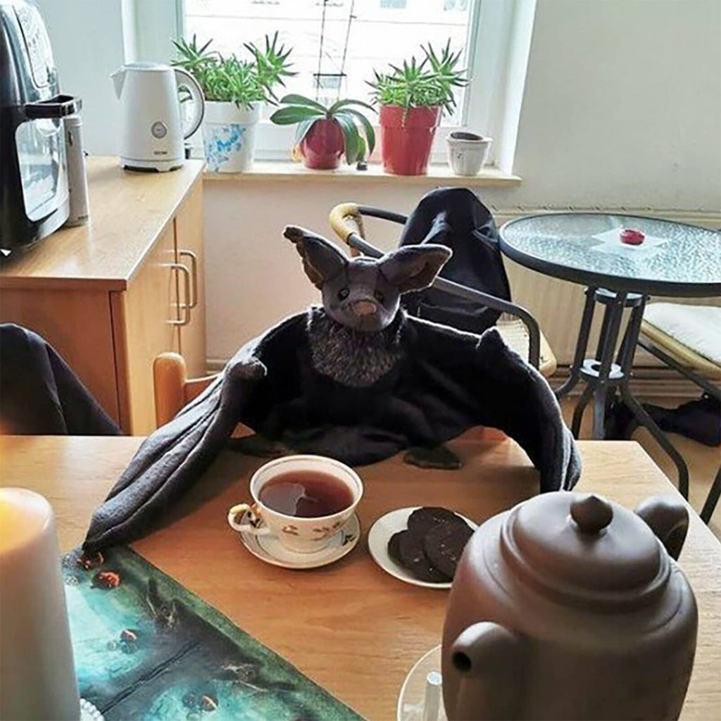 Plush bat with large wings sitting on a table with a cup of tea and cookies