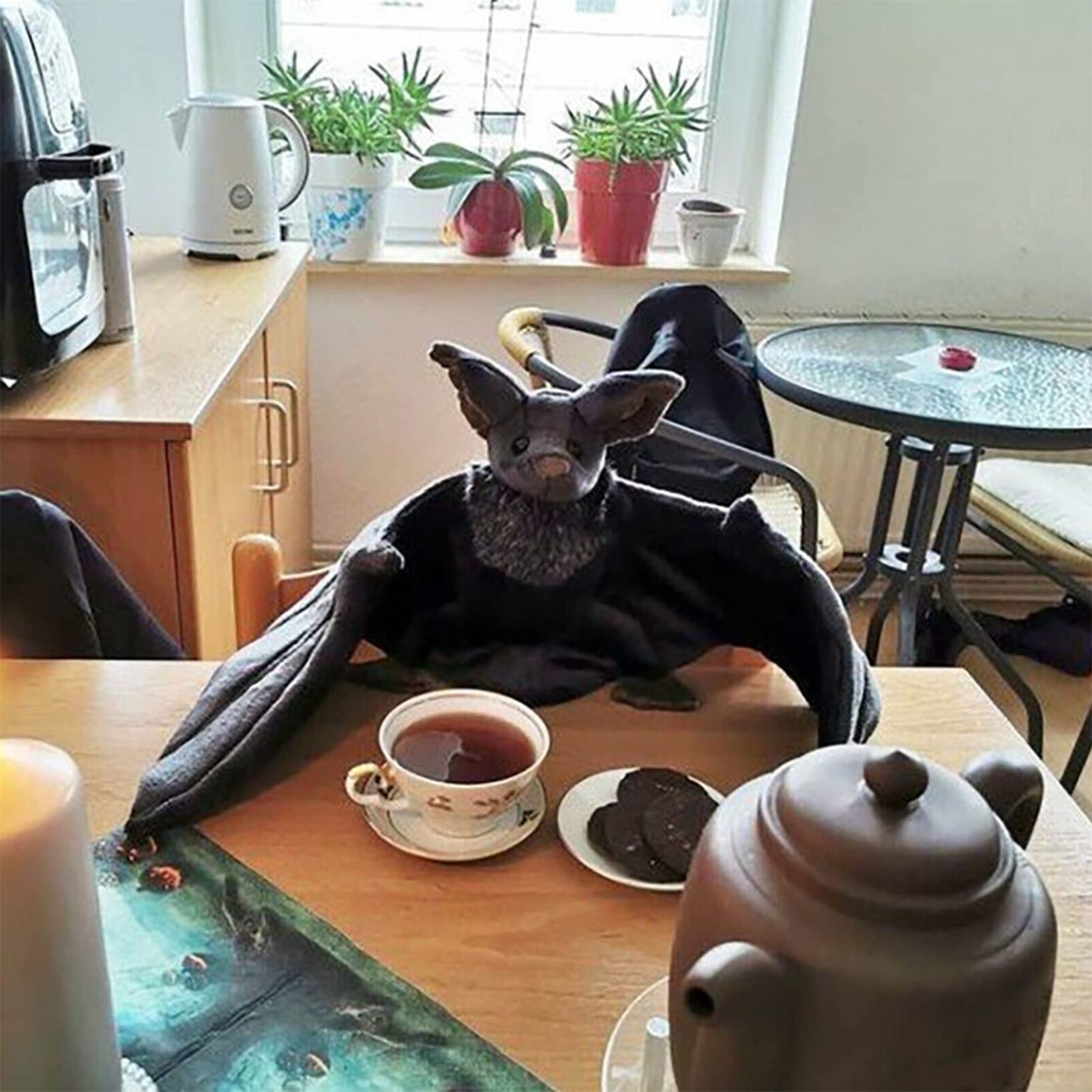 Plush bat with large wings sitting on a table with a cup of tea and cookies